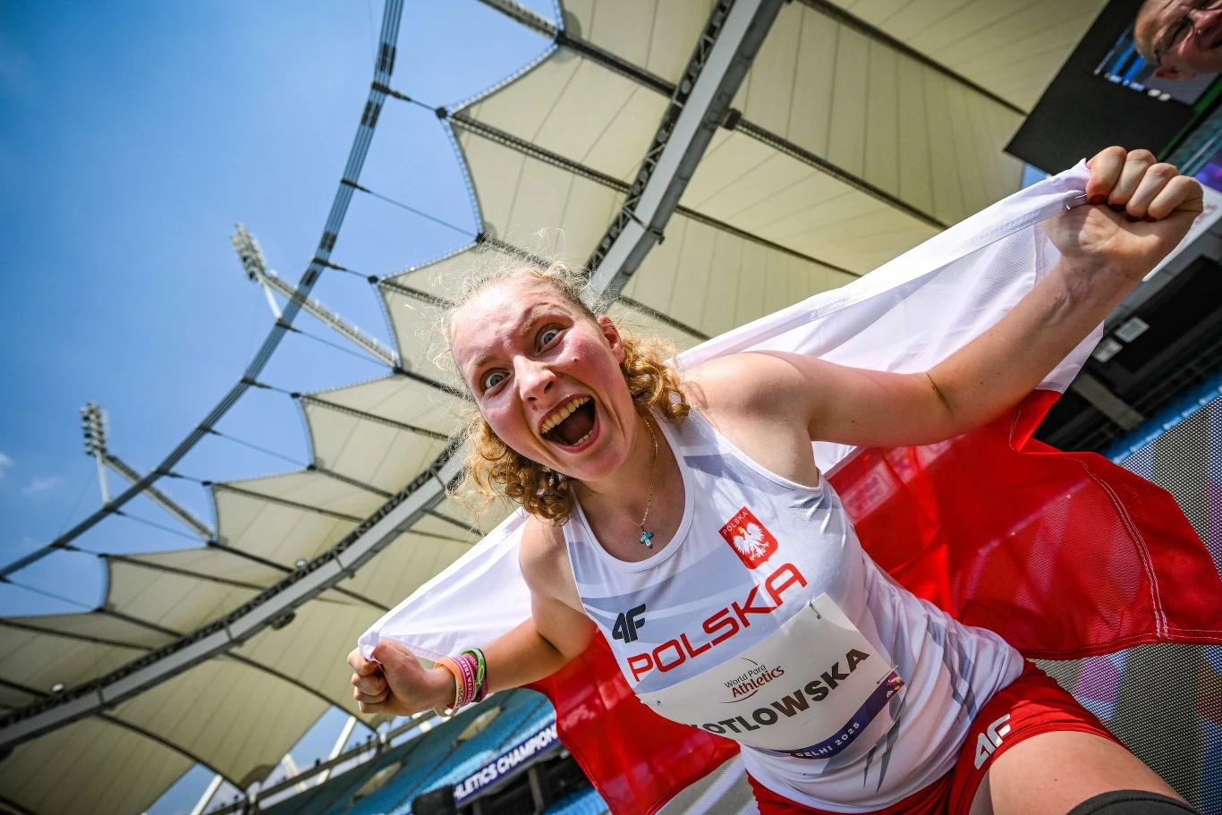 Faustyna Kotłowska, radość mistrzyni świata w paralekkoatletyce Szczęśliwa lekkoatletka z biało-czerwoną flagą Polski rozpościera flagę na stadionie, ubrana w sportowy strój z nazwiskiem i napisem Polska, w tle widoczne trybuny stadionu.