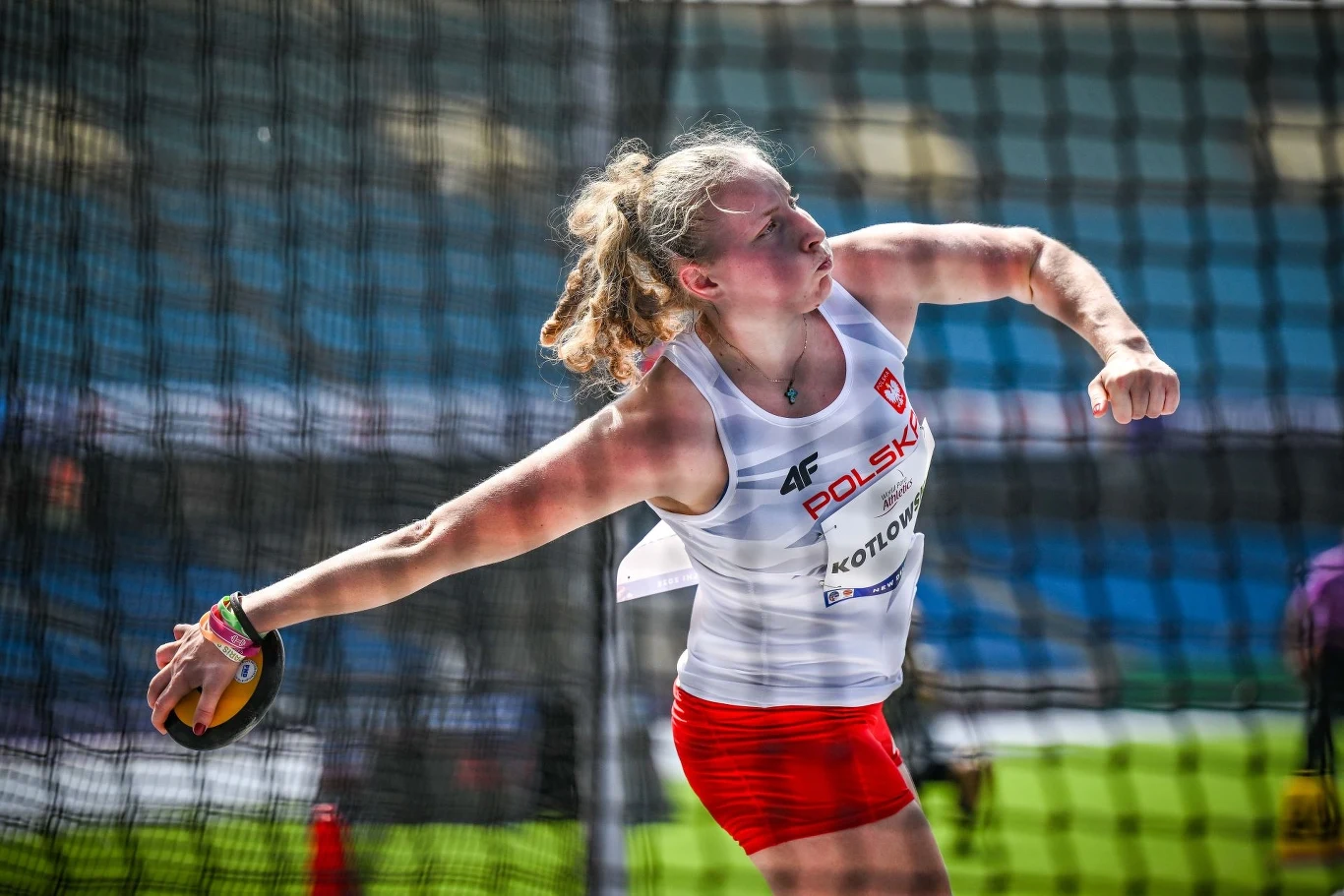 Faustyna Kotłowska w decydującej próbie Lekkoatletka w stroju reprezentacji Polski wykonuje rzut dyskiem na stadionie, z widoczną intensywnością sportowego wysiłku i skupieniem na twarzy.