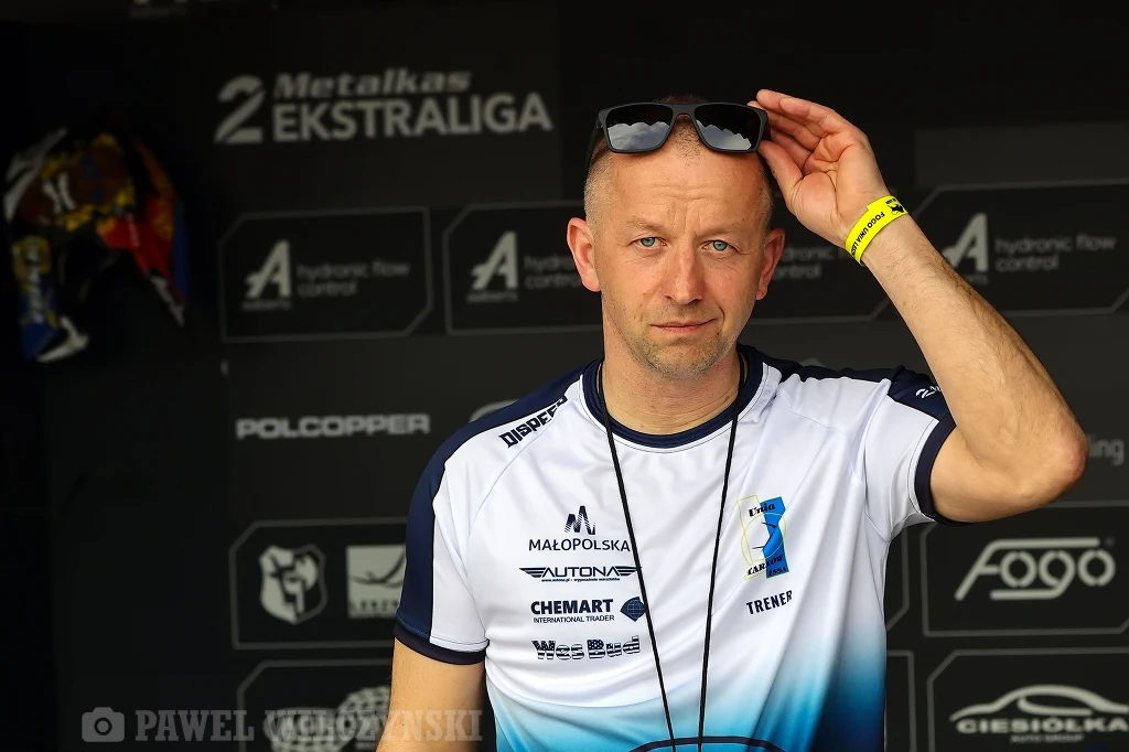 Trener Unii Tarnów Stanisław Burza Mężczyzna w sportowej koszulce trzyma okulary przeciwsłoneczne na głowie, w tle widoczne logotypy sponsorów i partnerów imprezy sportowej.