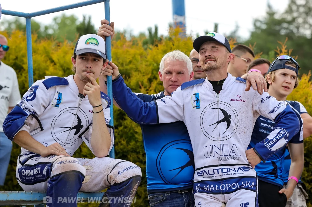 Grupa mężczyzn ubrana w sportowe stroje drużyny żużlowej Unia Tarnów, niektórzy mają na sobie czapki z daszkiem; w tle widoczna zieleń, atmosfera luźna i koleżeńska.