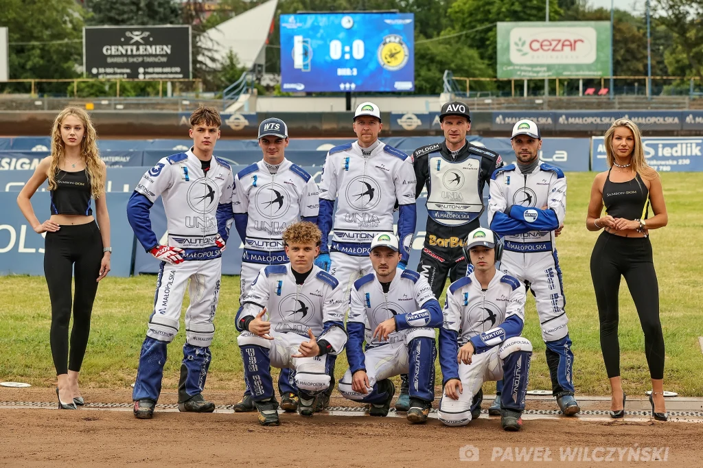 Zespół Unii Tarnów Grupa sportowców w białych i niebieskich strojach drużyny żużlowej pozuje na torze razem z dwiema kobietami ubranymi na czarno, widoczne tło stadionu oraz tablica wyników.