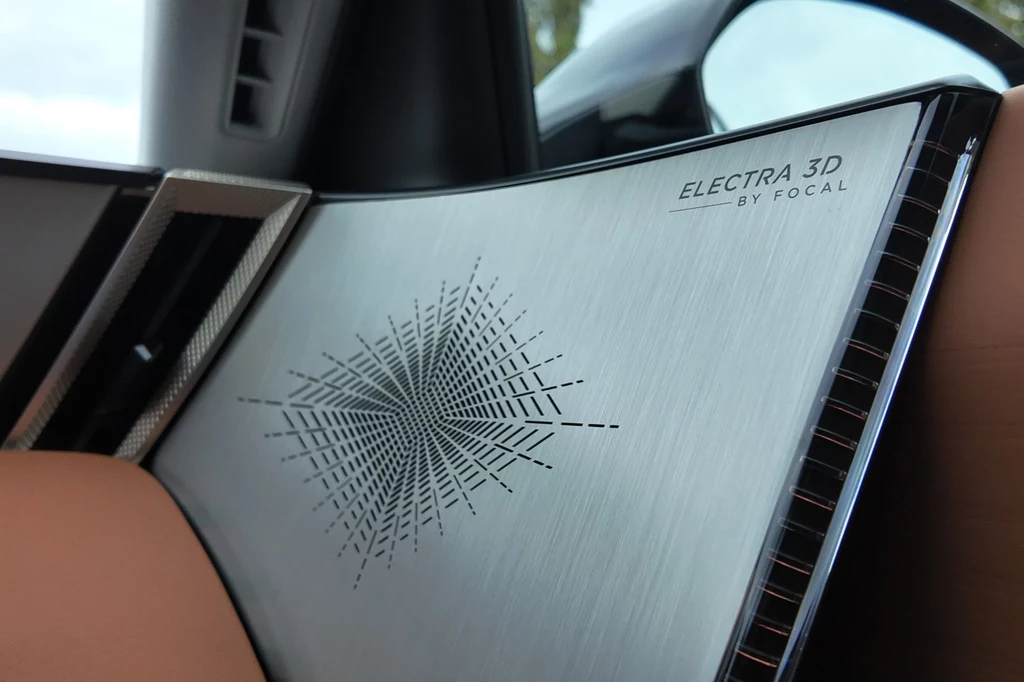Metaliczny panel głośnika samochodowego z napisem Electra 3D by Focal, geometryczny wzór wycięty laserowo, elementy wnętrza samochodu, w tle fragment drzwi oraz skórzane wykończenia.