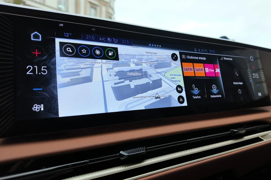 Panel multimedialny samochodu z widocznym ekranem dotykowym wyświetlającym system nawigacji, opcje klimatyzacji oraz menu multimediów, na ekranie wyświetlony jest szczegółowy plan miasta.