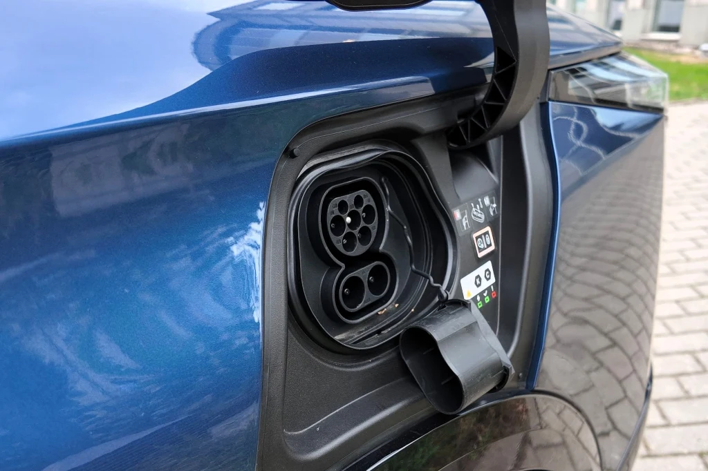 Gniazdo ładowania samochodu elektrycznego zamontowane w niebieskim pojeździe, widoczne ze zbliżenia, z otwartą klapką portu ładowania, na tle kostki brukowej.