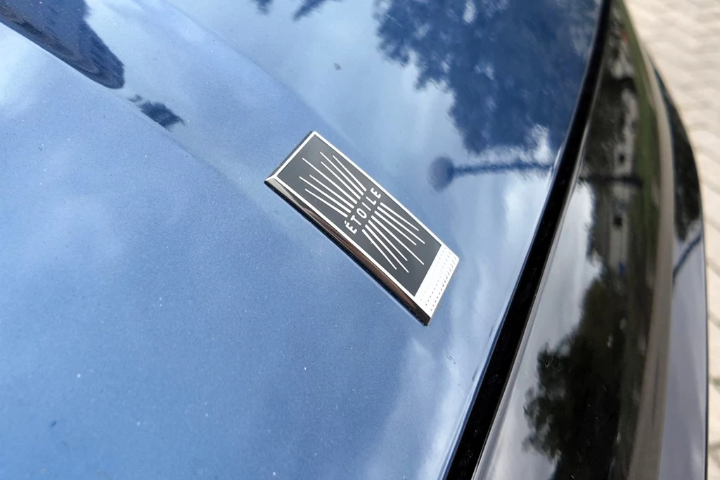 Emblemat z napisem Étoile umieszczony na masce ciemnoniebieskiego samochodu z odbiciem otoczenia na lakierze.