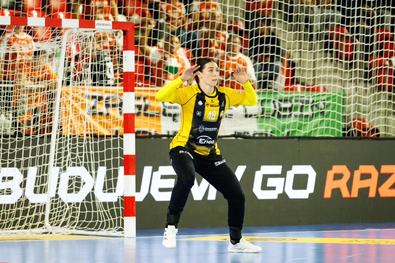 Zapowiedź 5. serii ORLEN Superligi Kobiet Bramkarz drużyny piłki ręcznej w żółto-czarnym stroju przygotowany do obrony rzutu, stojący na linii bramkowej podczas meczu, za nim widoczne siatka oraz transparenty kibiców.