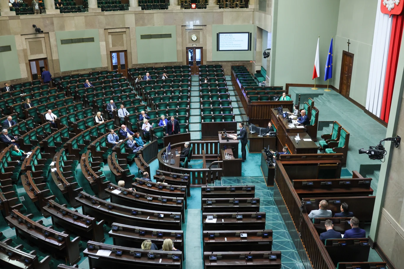 Najnowszy sondaż partyjny Sala obrad parlamentu z charakterystycznymi zielonymi ławami, pojedynczymi posłami siedzącymi w różnych miejscach oraz mównicą, przy której przemawia jedna osoba; w tle flagi Polski i Unii Europejskiej oraz ekran wyświetlający informacje