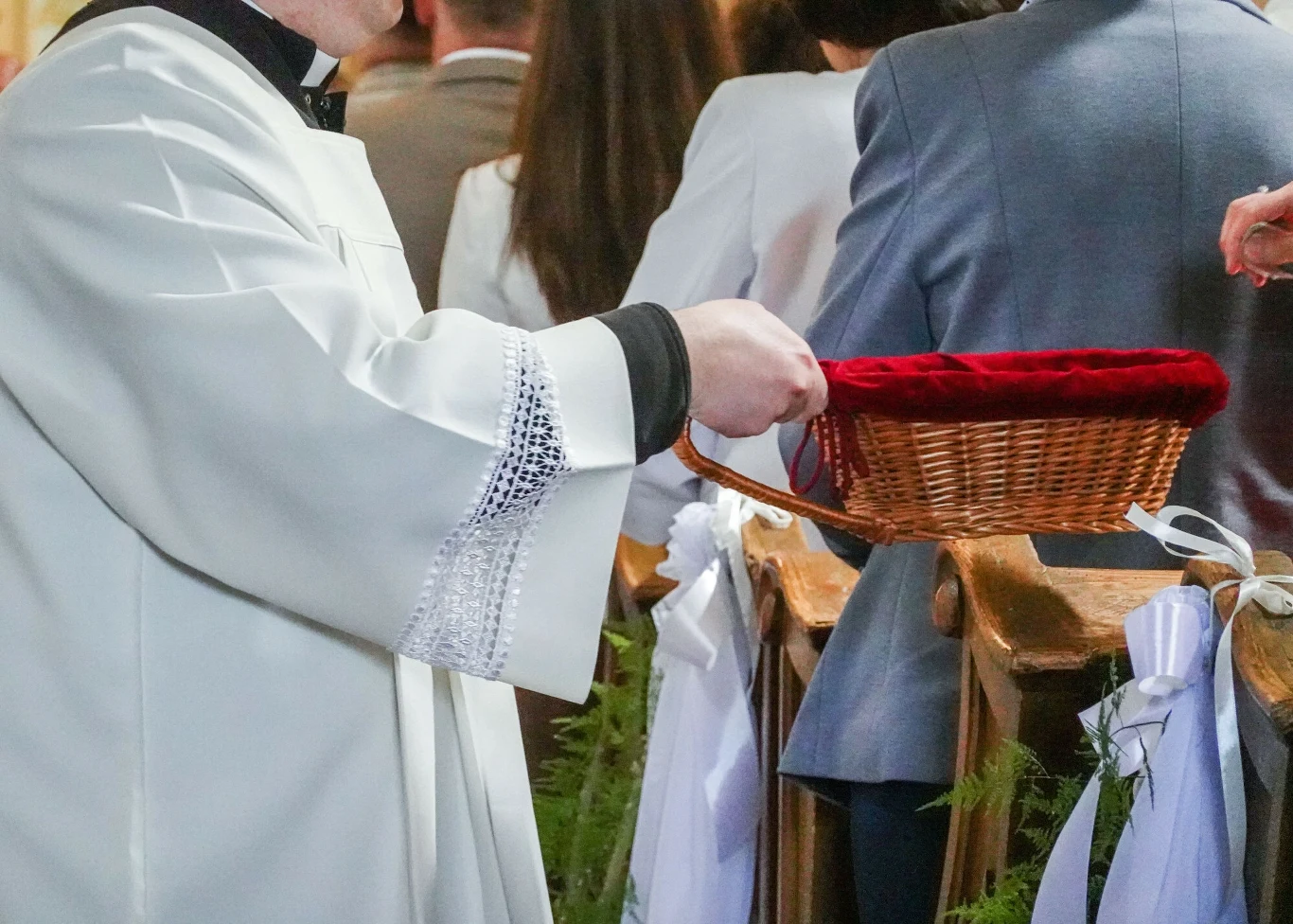 Pomysł podatku kościelnego jak dotąd nie znalazł rozwiązania prawnego w Polsce Ksiądz w białej szacie liturgicznej trzyma wiklinowy koszyczek przeznaczony na ofiarę pieniężną, w tle widoczne osoby siedzące w ławkach oraz eleganckie dekoracje kościelne.