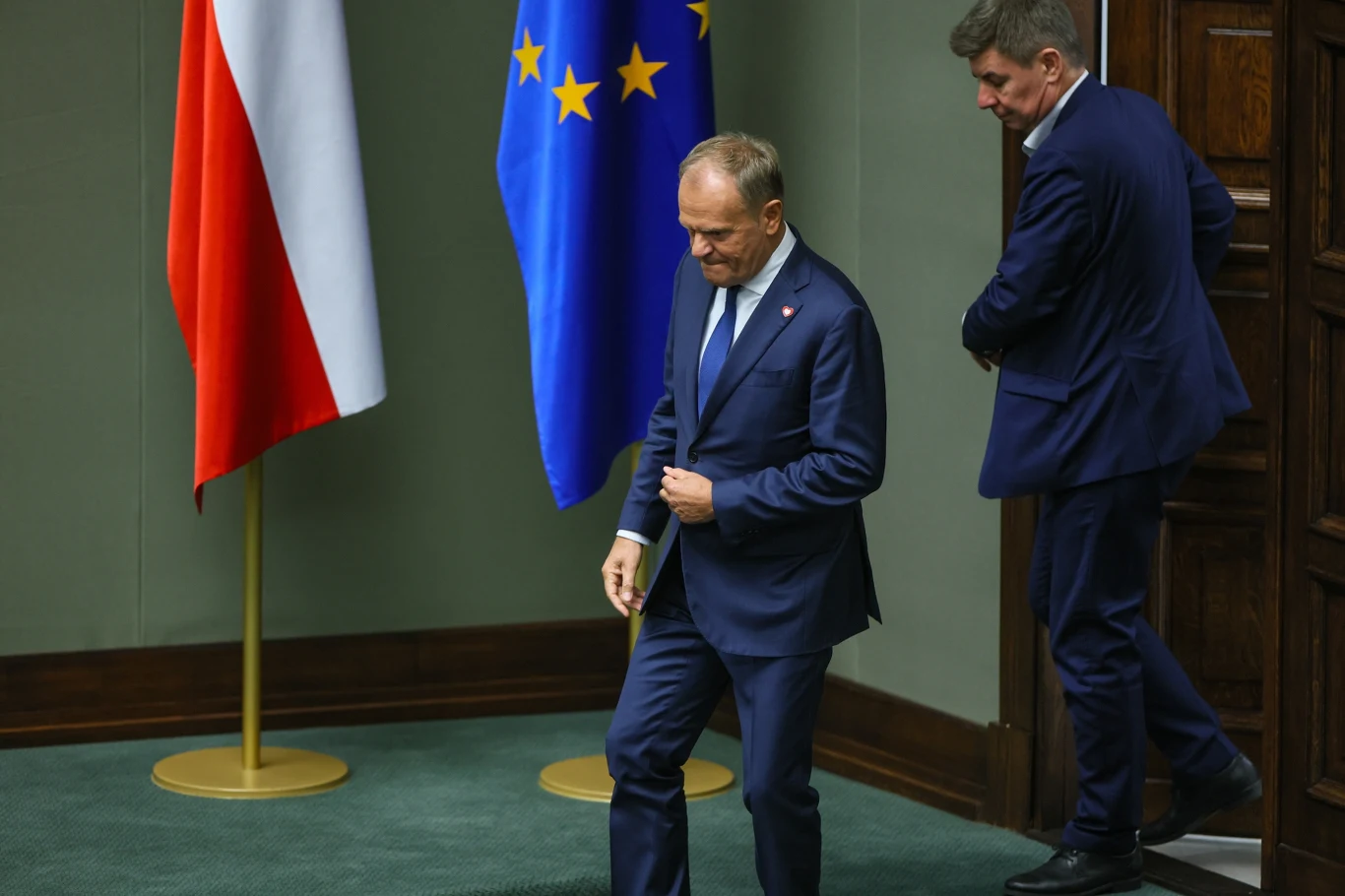 Premier Donald Tusk Dwóch mężczyzn w garniturach wychodzi z sali sejmowej, obok nich znajdują się flagi Polski oraz Unii Europejskiej.