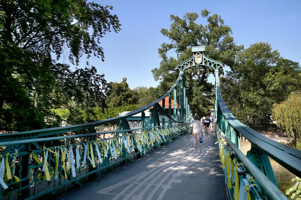Most z zielonej konstrukcji metalowej ozdobiony licznymi żółto-niebieskimi wstążkami, prowadzący przez park, po którym spacerują ludzie w słoneczny dzień.