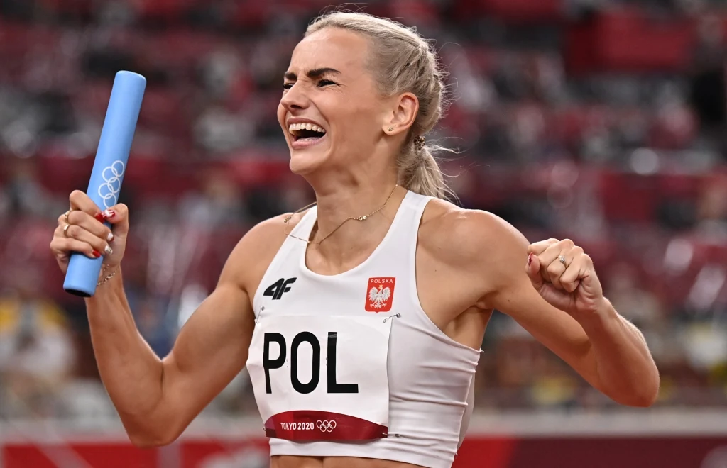 Szczęśliwa lekkoatletka w stroju z napisem POL trzyma niebieską pałeczkę sztafetową na tle stadionu podczas zawodów lekkoatletycznych.
