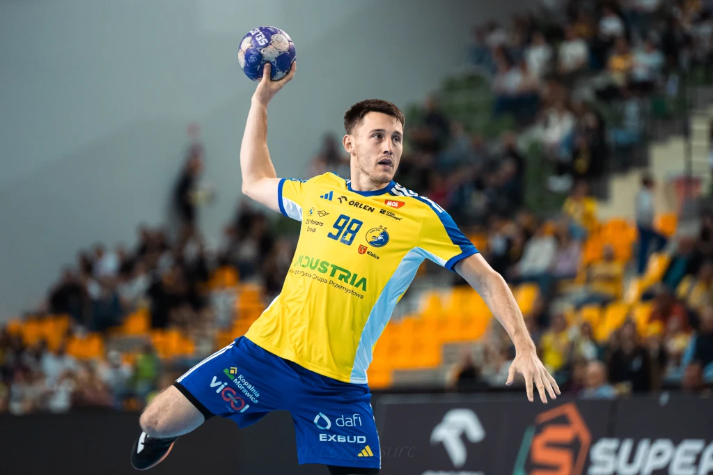 Podsumowanie 5. serii Orlen Superligi Zawodnik piłki ręcznej w żółto-niebieskim stroju unosi rękę z piłką w trakcie dynamicznej akcji na tle trybun z widownią.