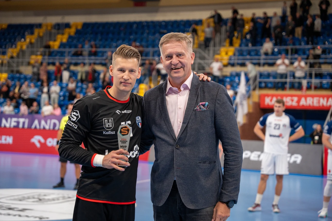 Zawodnik piłki ręcznej w czarnym stroju pozuje z nagrodą obok starszego mężczyzny w eleganckiej marynarce, na tle hali sportowej podczas meczu, w otoczeniu innych sportowców.