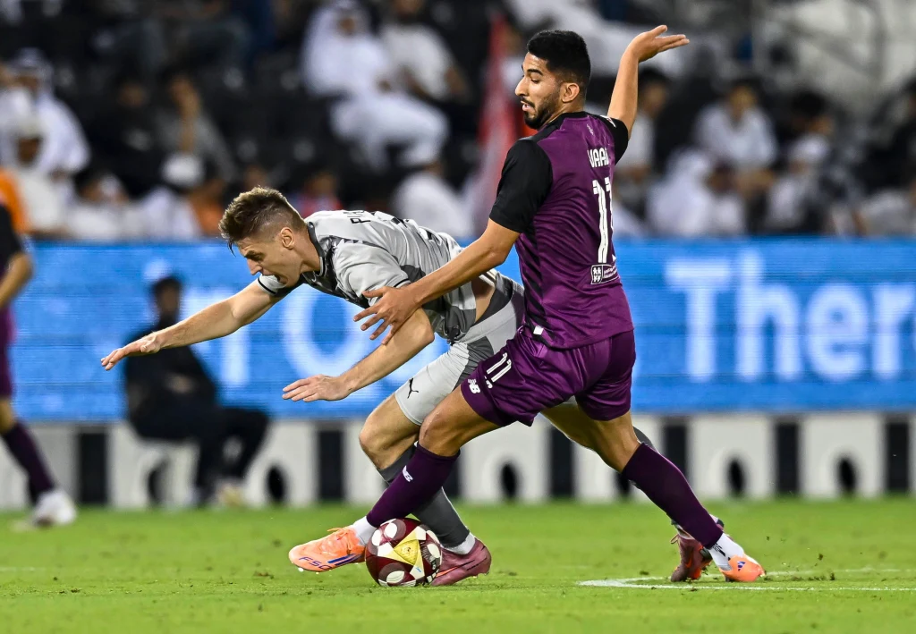 Dwóch piłkarzy rywalizuje o piłkę podczas meczu, jeden z nich upada na murawę starając się utrzymać równowagę, drugi stoi stabilnie, z wyciągniętym ramieniem. W tle widoczna publiczność oraz elektroniczne tablice reklamowe.