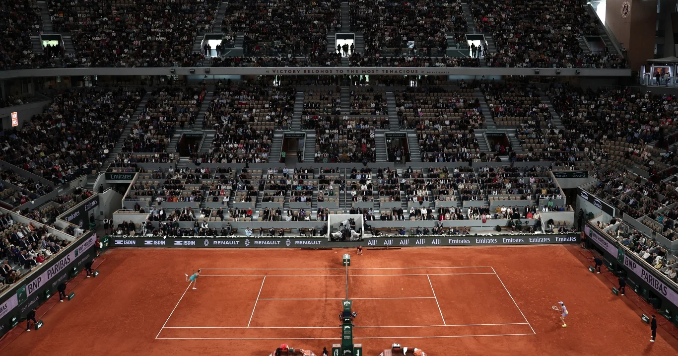 Centralny kort tenisowy podczas meczu, zawodnicy po przeciwnych stronach siatki, wokół wypełnione trybuny z liczną publicznością, ceglasta nawierzchnia kortu, światła stadionu.