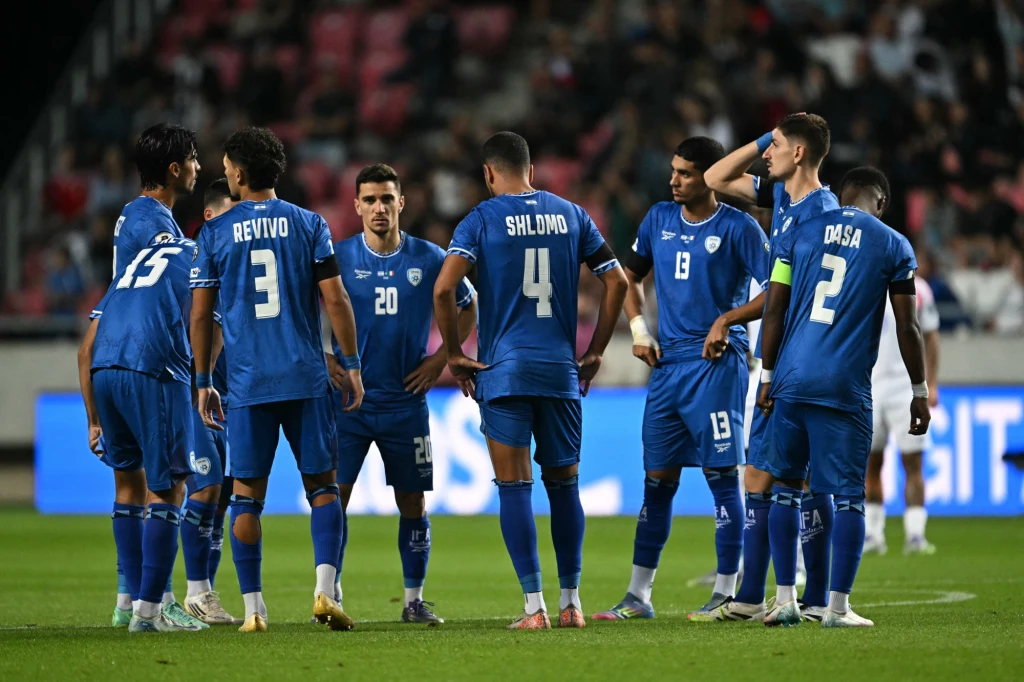 Reprezentacja Izraela Grupa piłkarzy w niebieskich strojach zgromadzona na murawie omawia strategię lub przygotowuje się do kolejnej akcji podczas meczu, w tle widoczni sędzia i przeciwnik.