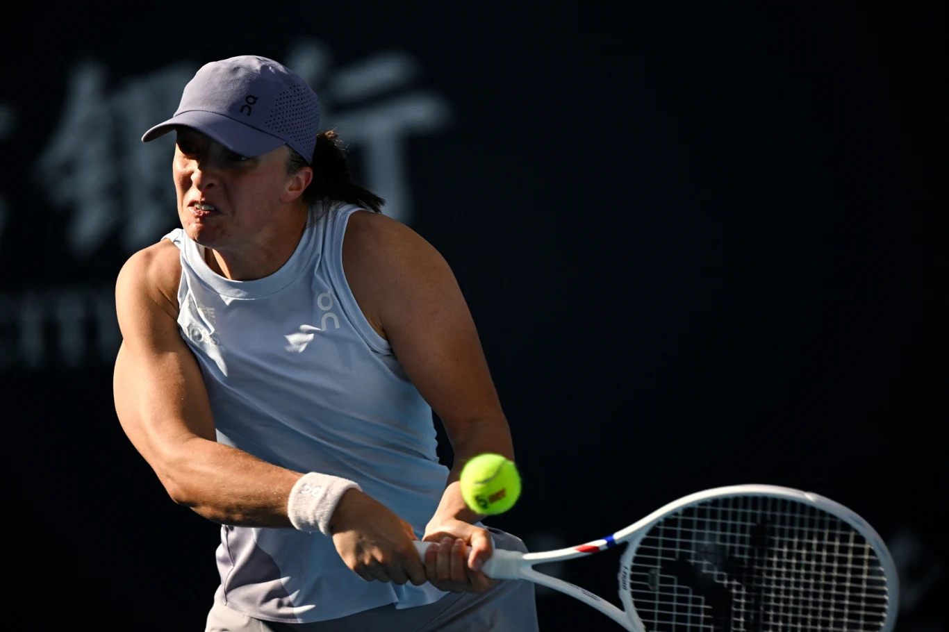 Tenisistka w białym stroju i czapce odbija piłkę rakietą, skupiona na grze, w tle ciemne rozmyte elementy.