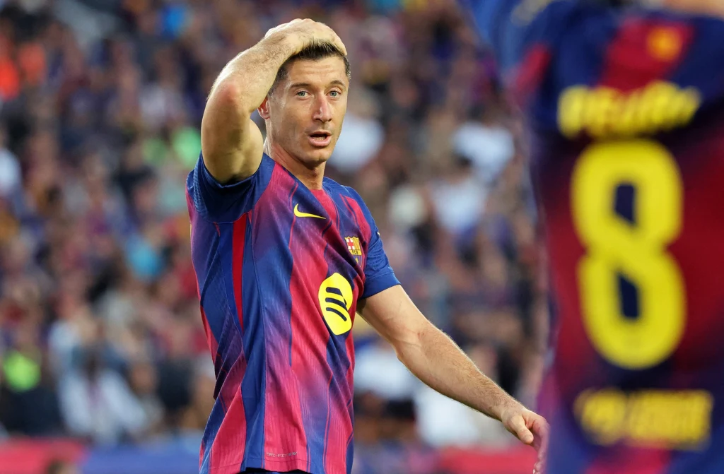 Robert Lewandowski Piłkarz w stroju FC Barcelony z numerem 9 na boisku, trzyma lewą rękę na głowie, na drugim planie fragment innego zawodnika w bordowo-granatowej koszulce.