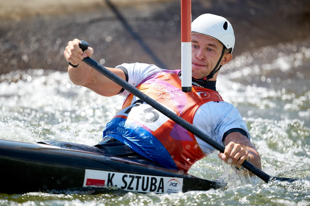 Kajakarz w kasku oraz kamizelce ochronnej pokonuje tor wodny, dynamicznie manewrując wśród przeszkód na wodzie, widoczny numer startowy oraz nazwisko zawodnika na kajaku.