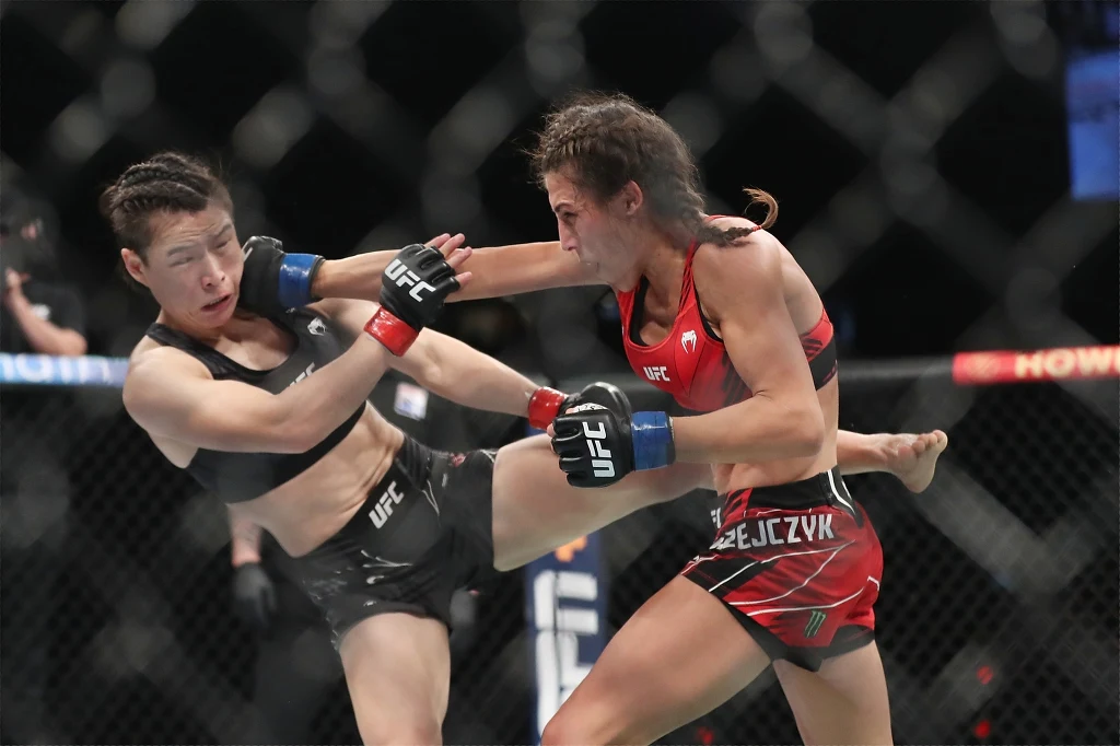 Dwie zawodniczki MMA wymieniają ciosy w oktagonie, jedna z nich zadaje kopnięcie nogą w tułów przeciwniczki, obie ubrane w sportowe stroje i rękawice, scena dynamiczna, w tle widać klatkę i elementy otoczenia areny walki.