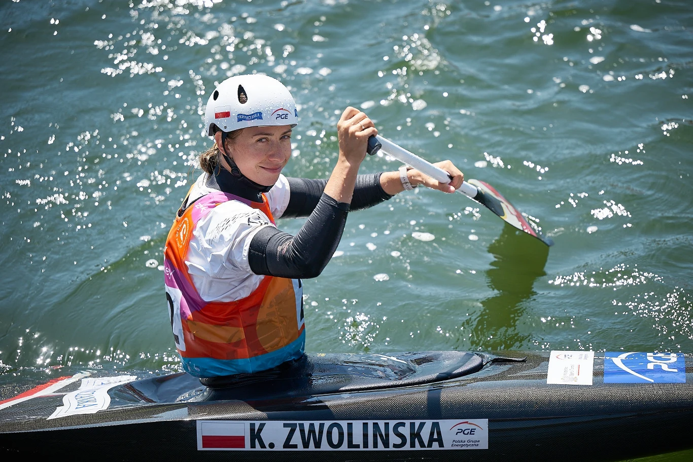 Sportsmenka w kajaku podczas zawodów, w kolorowej kamizelce asekuracyjnej i kasku, trzyma wiosło, wokół woda, widoczny napis z nazwiskiem K. Zwolińska na kajaku.