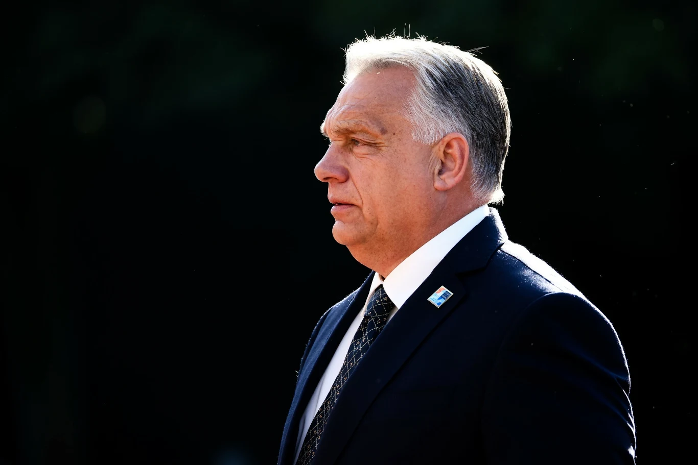 Premier Węgier Viktor Orban Mężczyzna w średnim wieku w garniturze, białej koszuli i krawacie stoi profilem na tle ciemnego, rozmytego tła, jego sylwetka jest dobrze oświetlona światłem dziennym.