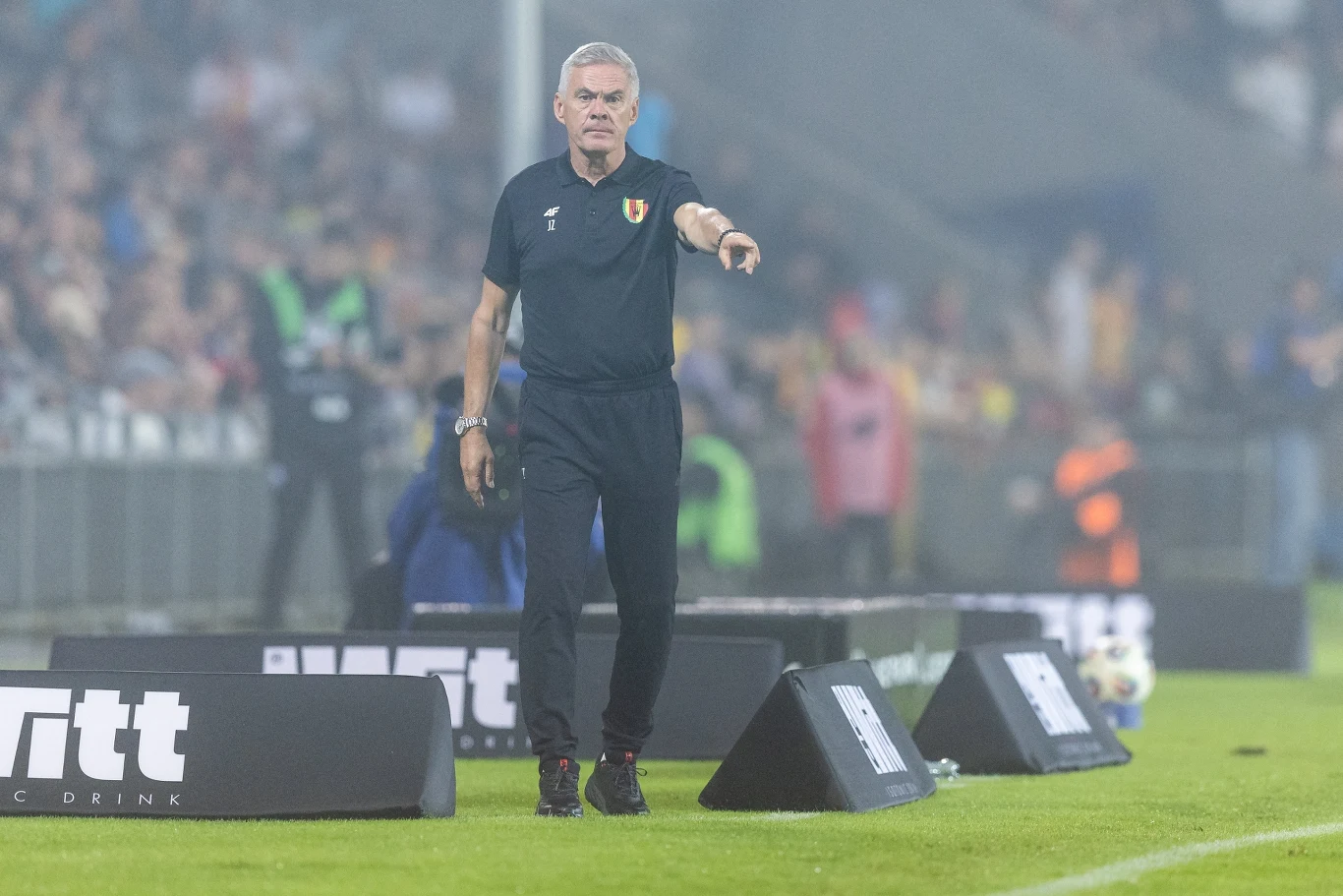 Trener piłkarski ubrany w czarny strój wskazuje palcem w kierunku boiska, stojąc przy linii bocznej podczas meczu, w tle widoczna rozmyta publiczność na trybunach.