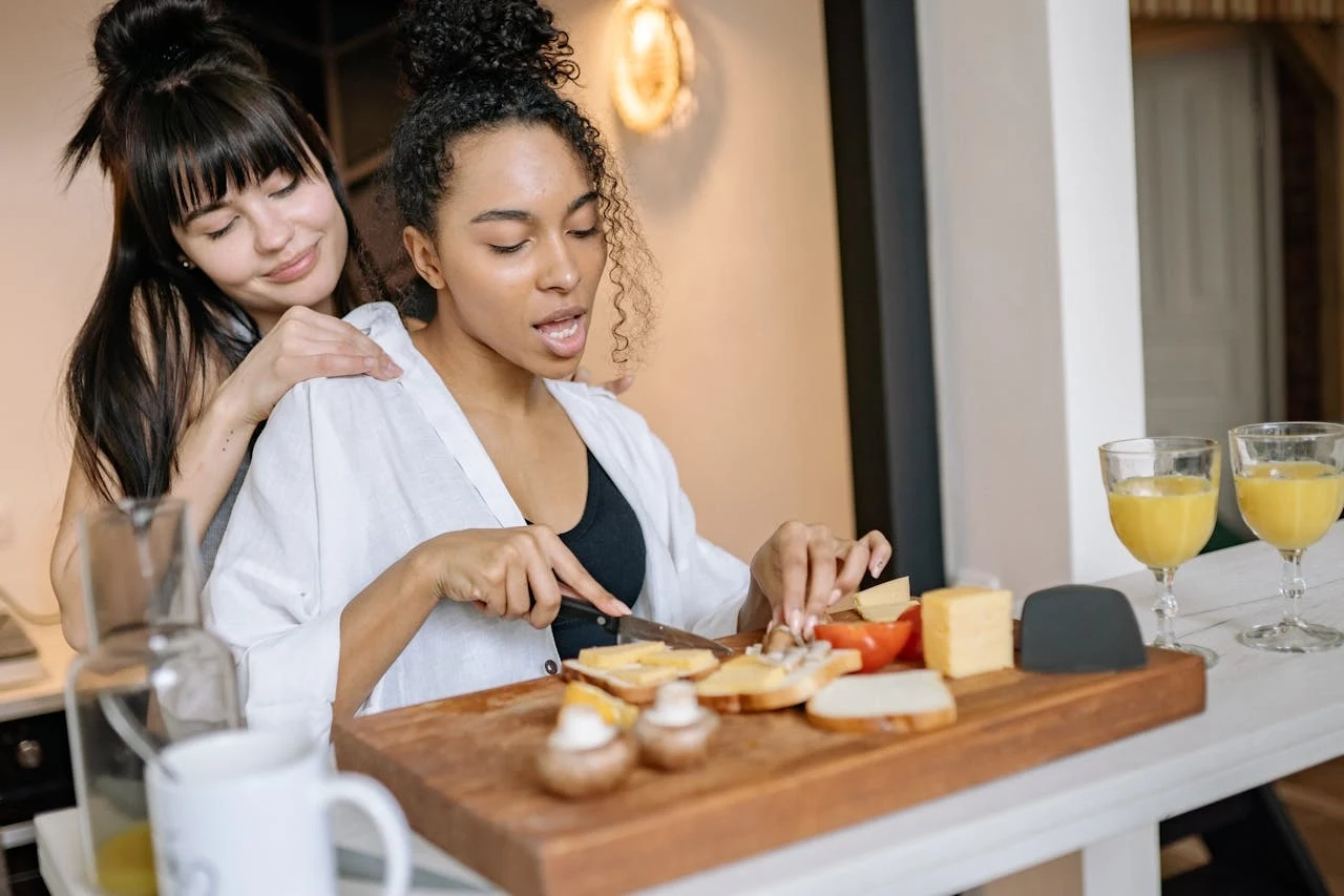 Dlaczego białko jest tak ważne w diecie kobiet? Dwie kobiety w kuchni przygotowują śniadanie, jedna kroi sery i warzywa na drewnianej desce, druga przytula ją od tyłu; na blacie stoją dwie szklanki z sokiem pomarańczowym oraz kawałki sera i pieczarki.