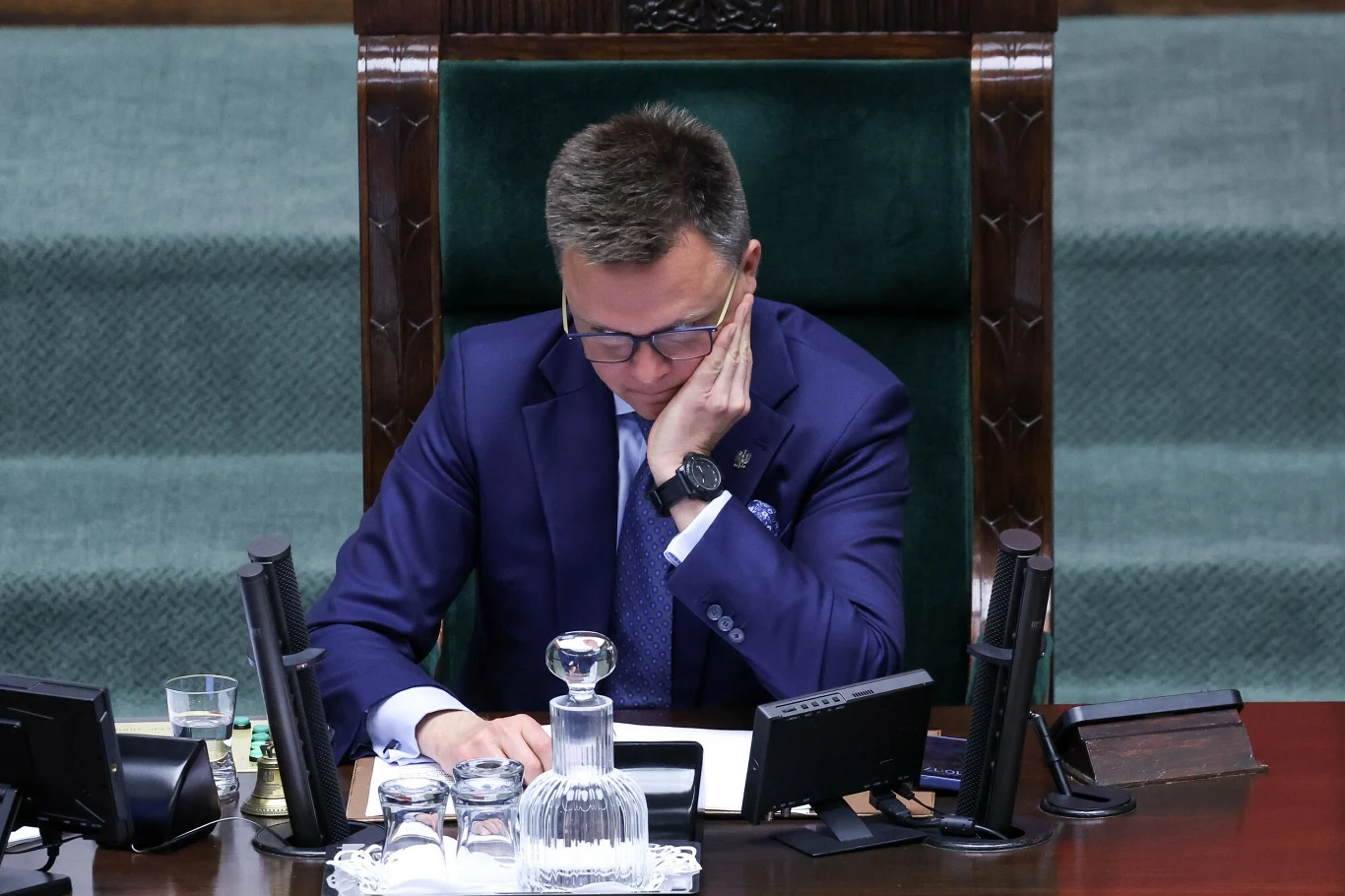 Marszałek Sejmu Szymon Hołownia Mężczyzna w garniturze i okularach siedzący za dużym stołem z zielonym obiciem, przed nim szklana karafka z wodą, szklanki oraz dokumenty, z tyłu wysoki ciemny fotel.