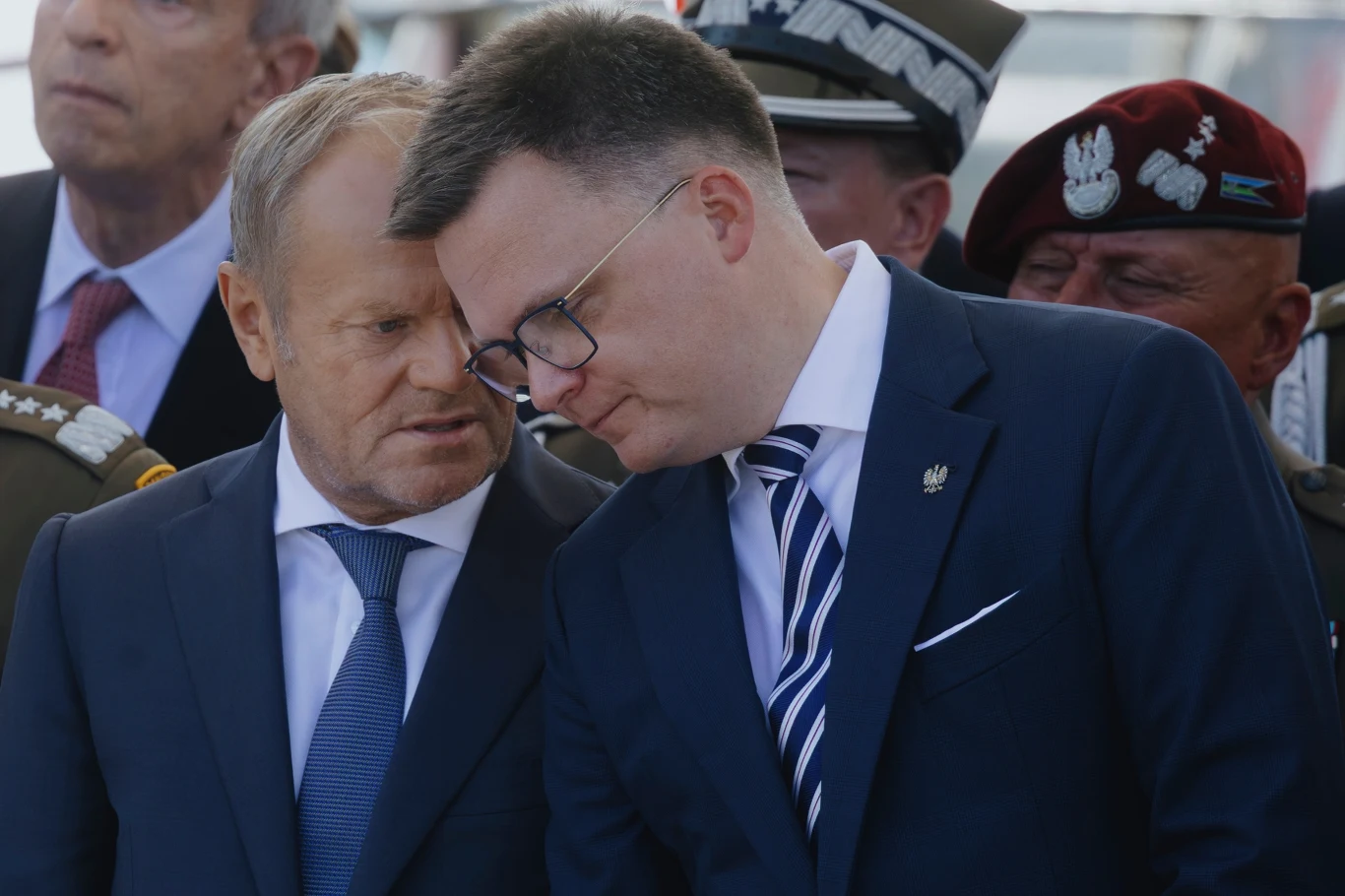 Dwóch mężczyzn w garniturach pochylonych ku sobie, prowadzących szeptaną rozmowę na tle mundurowych żołnierzy oraz oficjeli, co sugeruje poważny charakter spotkania.