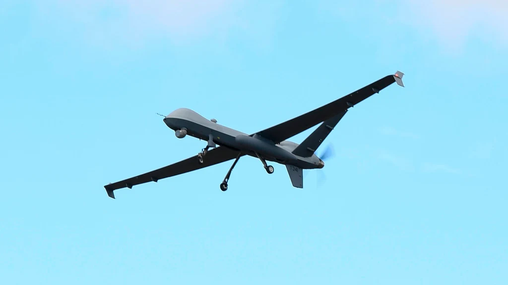 MQ-9 Reaper Wojskowy dron lecący na tle czystego, niebieskiego nieba, widoczny w locie z lekkiej perspektywy od dołu, wyraźnie widoczne są jego długie skrzydła i wirujące śmigło.