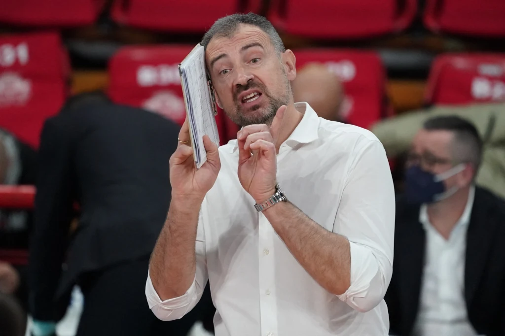 Mężczyzna w białej koszuli trzyma notatnik i wykonuje gest ręką, prawdopodobnie wyjaśniając coś lub instruując podczas wydarzenia sportowego, w tle zarysowani inni ludzie oraz czerwone siedzenia.