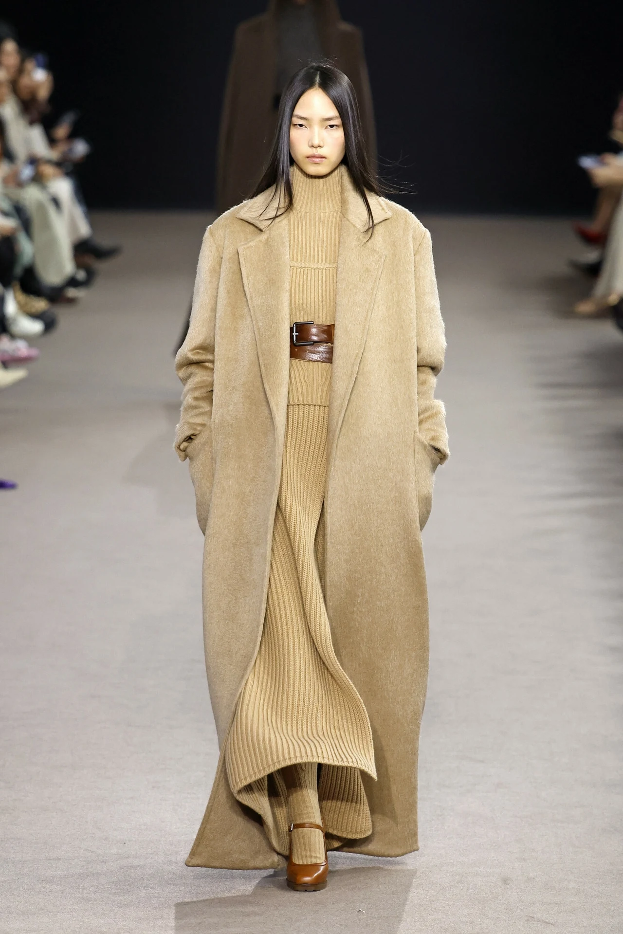 Pokaz Max Mara na jesień-zimę 2024/2025 Modelka idzie po wybiegu ubrana w beżowy dzianinowy komplet składający się z długiego płaszcza, swetra z golfem oraz spódnicy, przepasana szerokim brązowym paskiem; na nogach brązowe buty na obcasie, włosy proste, luźno opadające.