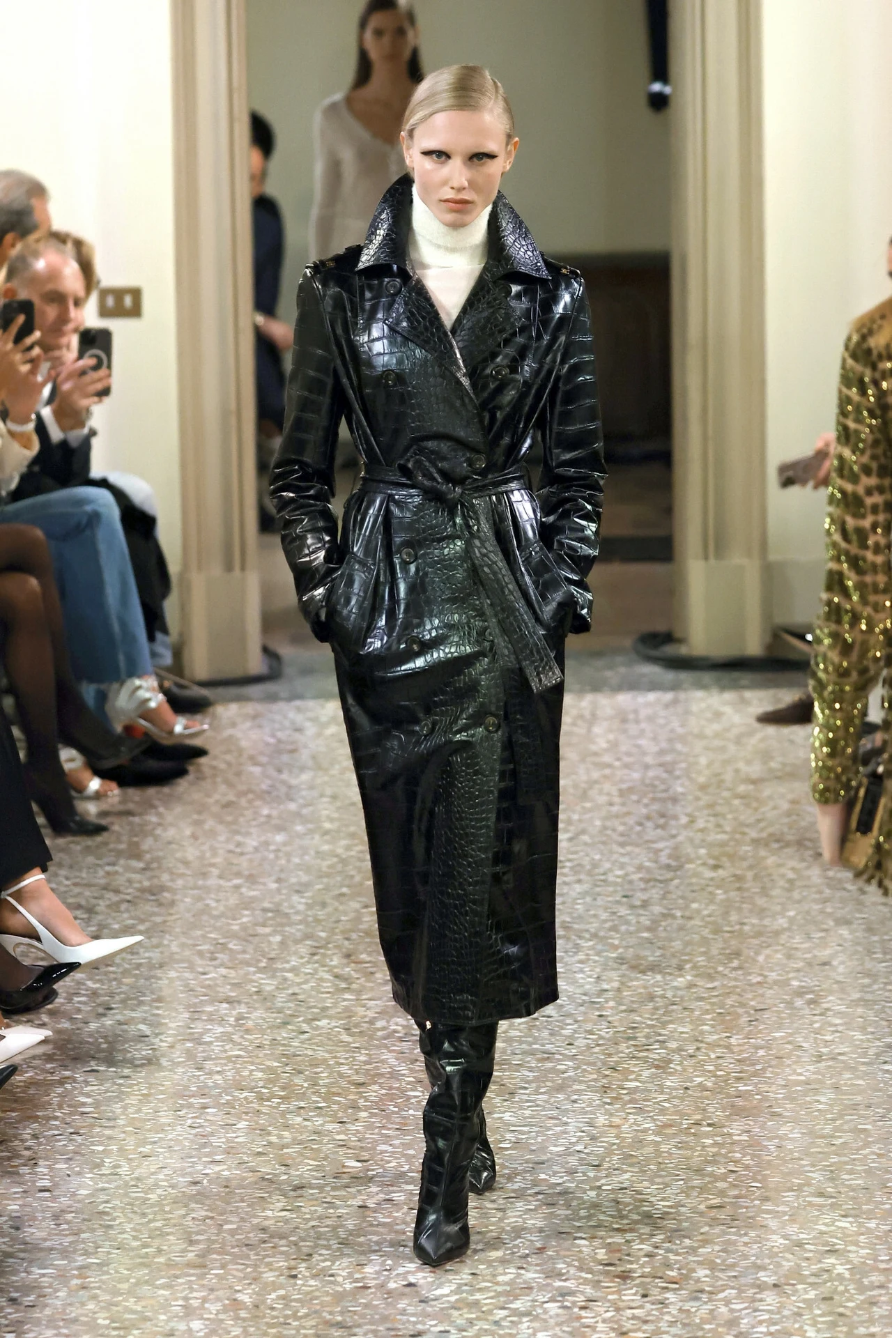 Pokaz Elisabetta Franchi jesień-zima 2024/2025 Modelka idzie po wybiegu w długim, czarnym, lakierowanym płaszczu z połyskującego materiału, pod spodem ma jasny golf, a jej włosy są gładko zaczesane; wokół siedzi publiczność i fotografowie.