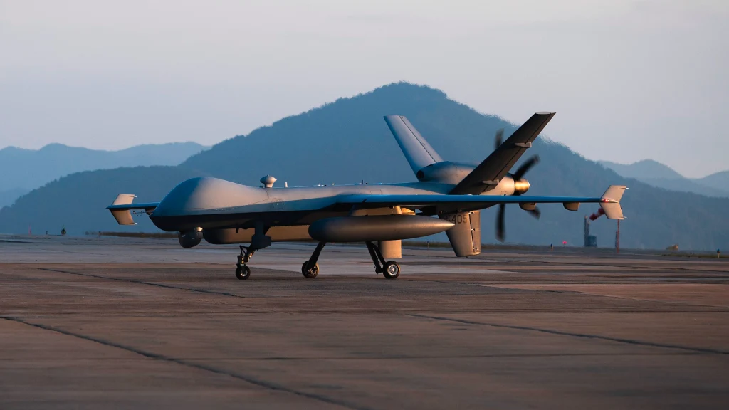 Amerykanie rozmieszczają duże drony MQ-9 Reaper na zapalnym półwyspie Wojskowy dron o aerodynamicznej sylwetce stoi na płycie lotniska na tle gór i lekko zamglonego nieba, przygotowany do startu lub po lądowaniu, dominujące kolory to odcienie szarości i błękitu.