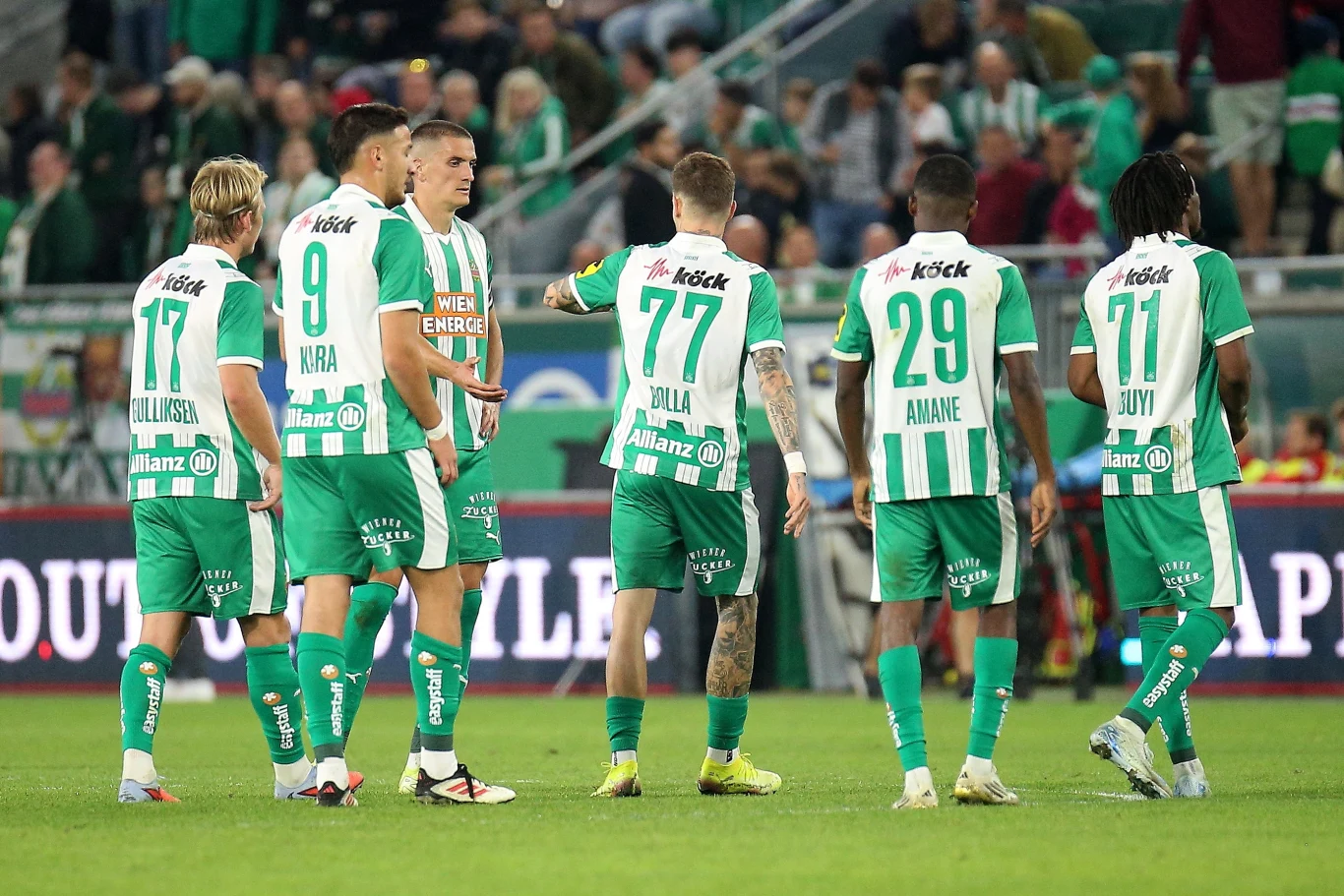 Piłkarze Rapidu przegrali derby Wiednia z Austrią aż 1:3 Grupa piłkarzy w zielono-białych strojach drużyny klubowej na stadionie piłkarskim, zawodnicy odwróceni częściowo plecami rozmawiają podczas meczu, w tle kibice na trybunach