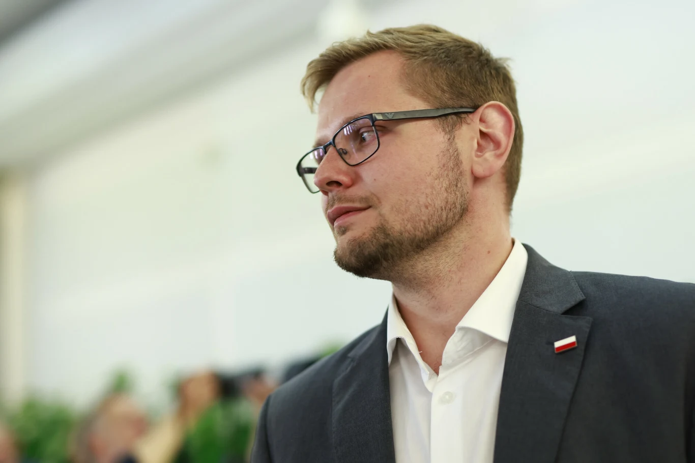 Michał Woś został wezwany do prokuratury Mężczyzna w garniturze z brodą i okularami, na klapie marynarki przypięta biało-czerwona polska flaga, rozmyte tło z postaciami i roślinami nadaje zdjęciu formalny charakter.