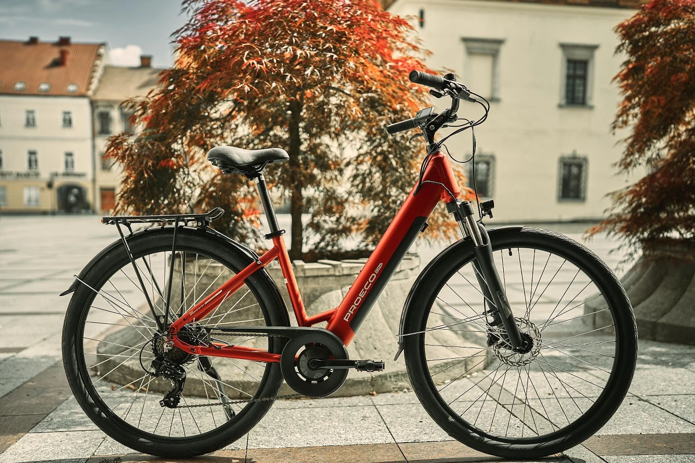 Zdjęcie główne przedstawiające rower elektryczny Czerwony rower elektryczny stoi na tle miejskiego placu, otoczony jesiennymi drzewami o rudych liściach, w tle widoczne są zabytkowe budynki.