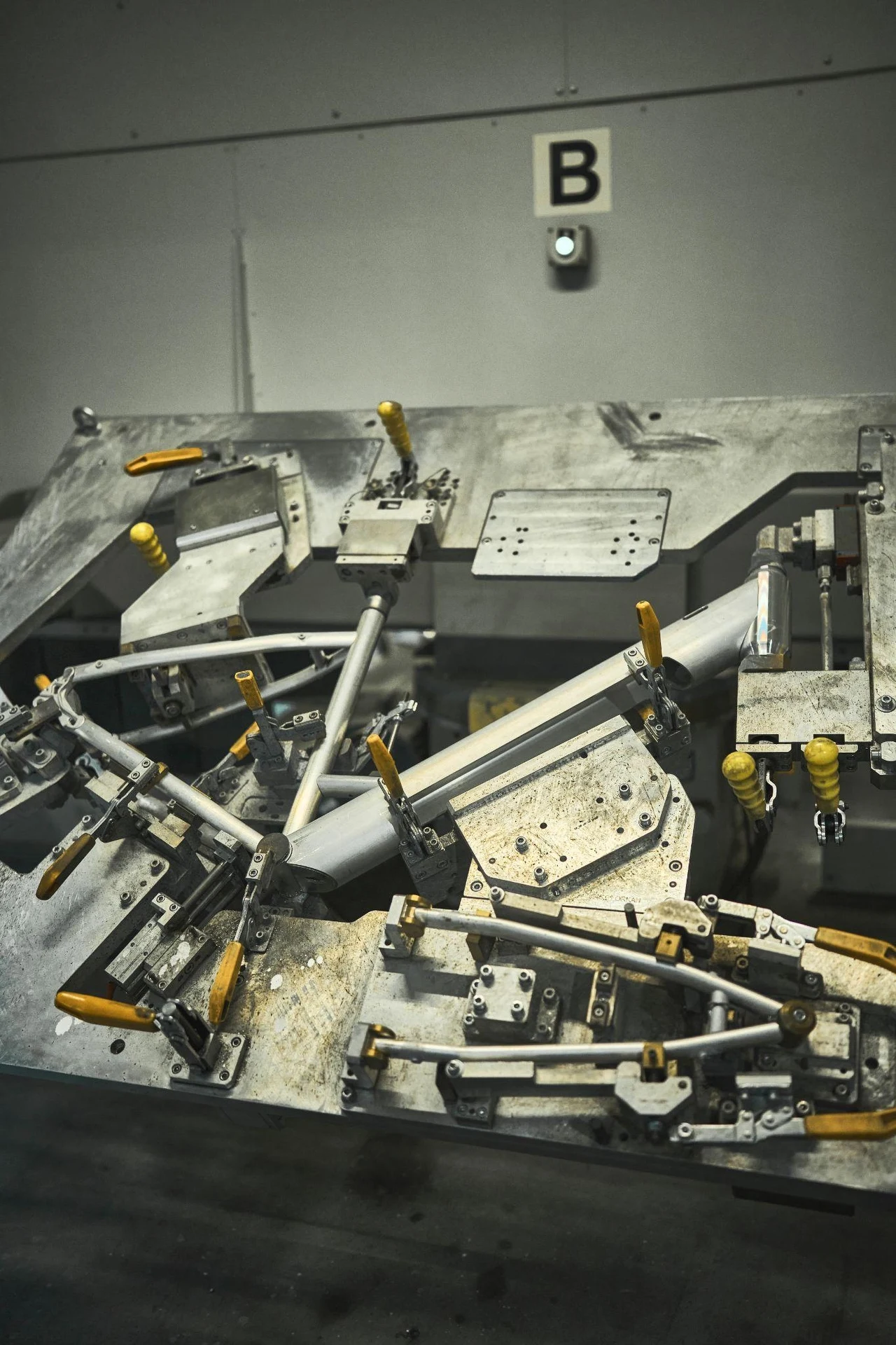 Produkcja ram do rowerów Metalowy stół montażowy z zamontowanymi elementami mocującymi, pokrętłami i uchwytami w jasnym, przemysłowym otoczeniu, na ścianie znajduje się oznaczenie literą B.
