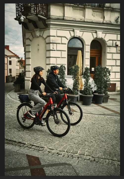 Ludzie jeżdżący na rowerach elektrycznych Dwoje rowerzystów w kaskach rowerowych przemierza brukowaną uliczkę w centrum miasta, mijając zabytkową kamienicę otoczoną roślinami w dużych donicach.