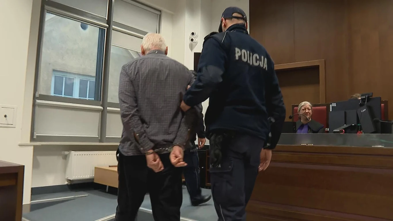 Mężczyzna skazany na 15 lat więzienia. Zbrodnię popełnił 30 lat temu Policjant prowadzi starszego mężczyznę w kajdankach przez salę sądową, w tle sędzia siedzący za stołem sędziowskim i druga osoba przy komputerze.