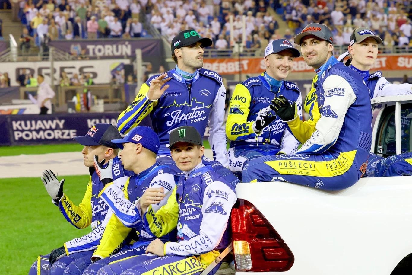 Grupa żużlowców w niebiesko-żółtych kombinezonach sportowych siedzi na białym samochodzie podczas prezentacji na stadionie żużlowym, w tle publiczność i banery reklamowe.