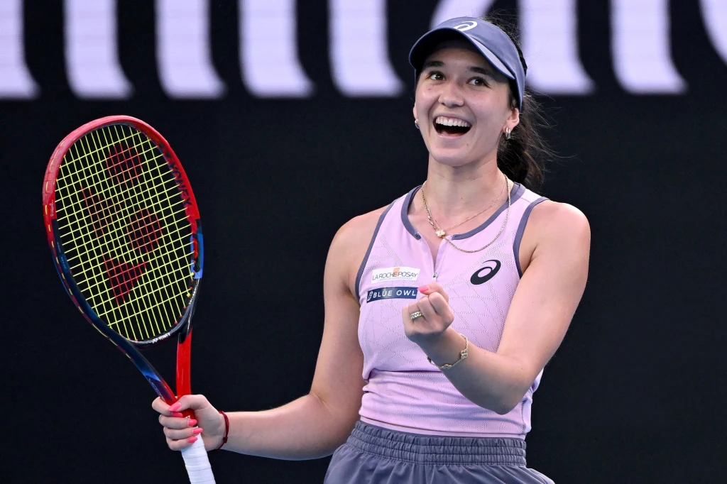 Eva Lys tenisistka w sportowej koszulce i czapce trzymająca rakietę tenisową oraz nagrodę, szeroko uśmiechnięta na tle kortu, wyrażając radość po meczu