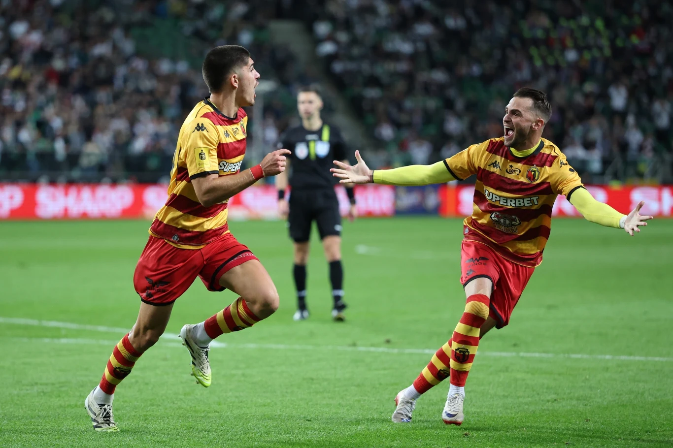 Jagiellonia Białystok zacznie udział w Lidze Konferencji od meczu z Hamrun Spartans Dwóch piłkarzy w żółto-czerwonych strojach zespołu świętuje zdobycie bramki na boisku, w tle widoczny sędzia oraz rozmyta publiczność na trybunach.