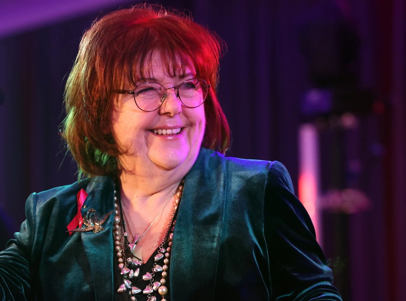 Maggie Reilly wystąpi w Polsce 30 września i 1 października Starsza kobieta w eleganckim, ciemnozielonym żakiecie z okularami na twarzy i szerokim uśmiechem, ma na sobie kilka naszyjników, siedzi w jasnym, kolorowym świetle, na tle ciemnych kotar.