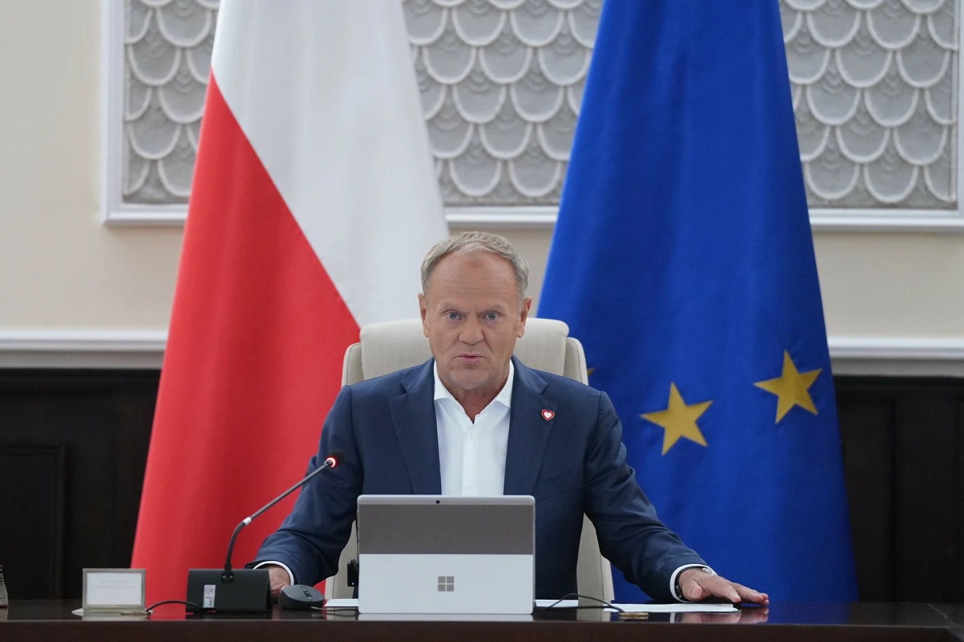 Donald Tusk popiera projekt podwyżki CIT da banków Mężczyzna w garniturze siedzący przy nowoczesnym biurku z laptopem, w tle widoczne flagi Polski oraz Unii Europejskiej, oficjalna atmosfera spotkania rządowego.
