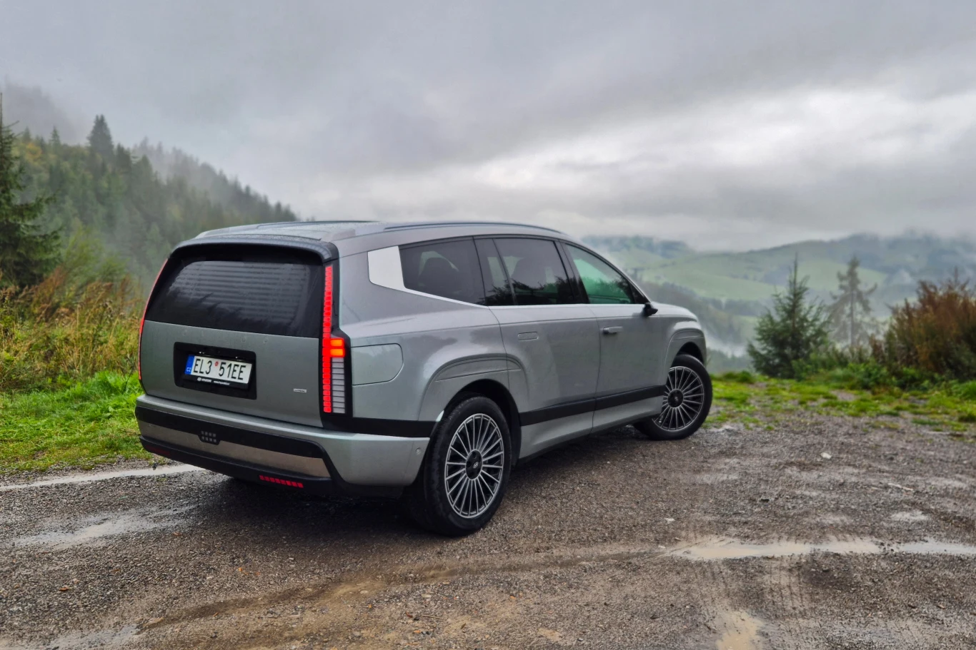 Srebrny SUV o nowoczesnej sylwetce zaparkowany na żwirowej nawierzchni, w tle rozciągające się górskie krajobrazy z mgłą i drzewami tworzącymi naturalny, spokojny klimat.