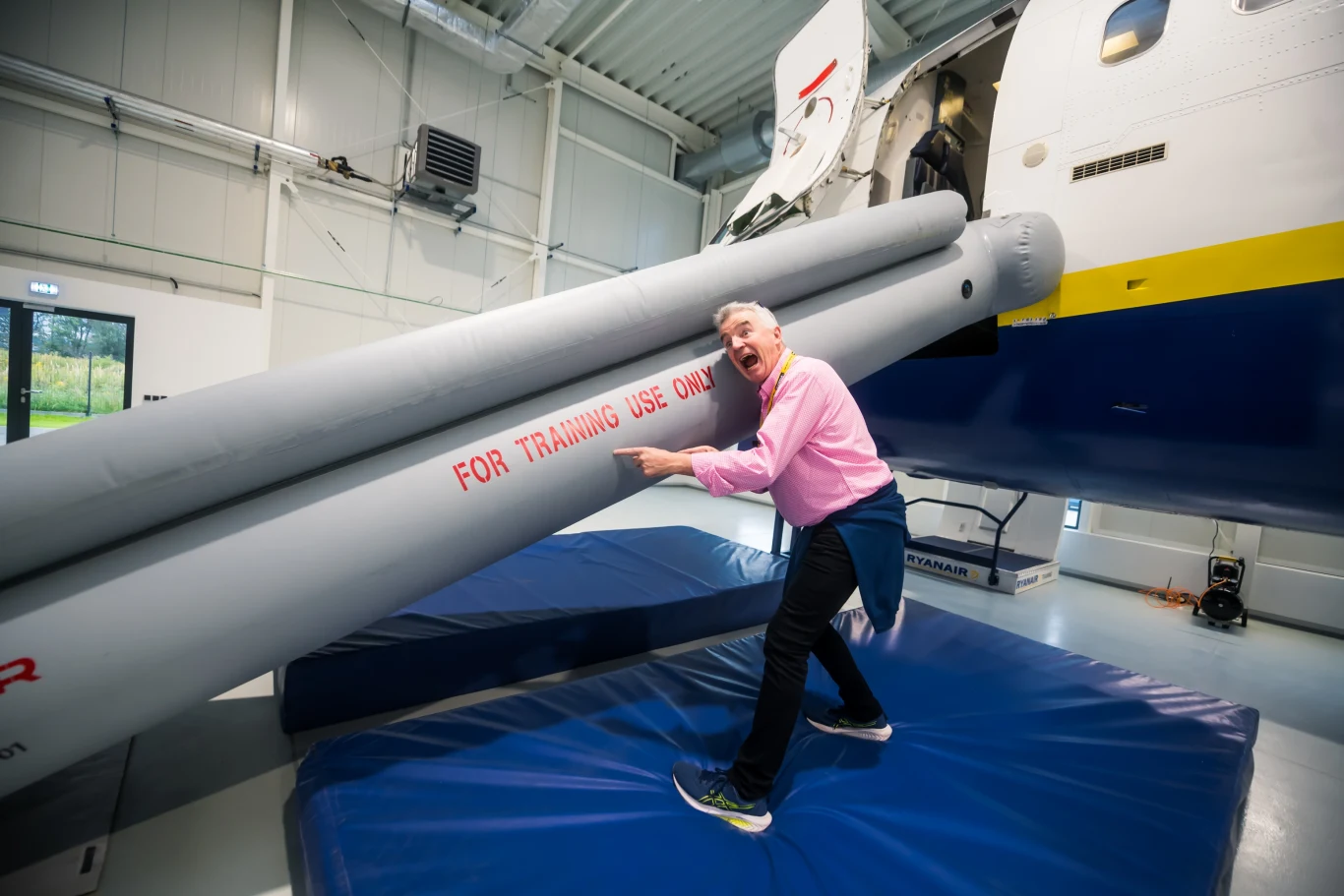 Prezes Ryanair Michael O'Leary prezentuje specjalistyczny kadłub treningowy. Mężczyzna przytulający duży, nadmuchiwany zjazd ewakuacyjny samolotu, znajdujący się wewnątrz hali treningowej, na niebieskich matach, urządzenie oznaczone napisem 'FOR TRAINING USE ONLY'.