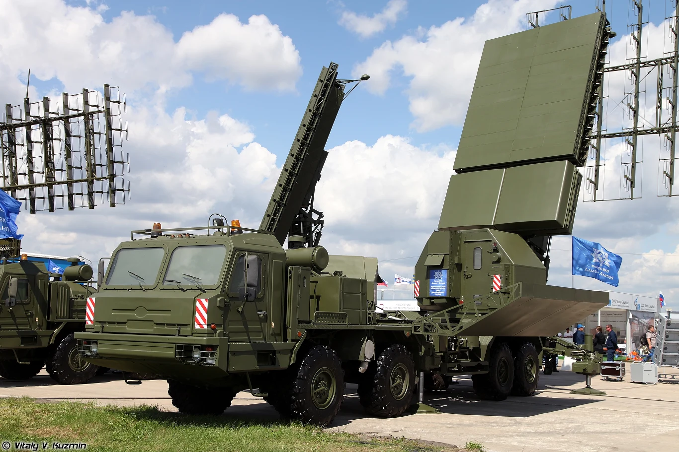 Wchodzący w skład systemu radarowego Niebo-M systemu S-400, pracujący w paśmie X radar RLM-S Wojskowy pojazd ciężarowy wyposażony w duży radar i maszty antenowe ustawiony na otwartym terenie, w tle widoczne są inne elementy wojskowej infrastruktury oraz niebo z chmurami.