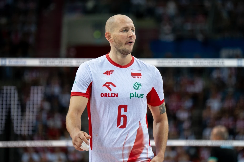 Bartosz Kurek Siatkarz w oficjalnym stroju reprezentacji Polski podczas meczu, w tle rozmyte sylwetki kibiców i fragment siatki.